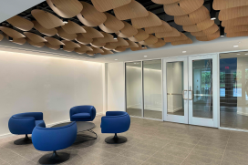 The lobby of an office building. Four blue chairs sit around a table. The decorative ceiling has wooden panels that look like scales or petals. 
