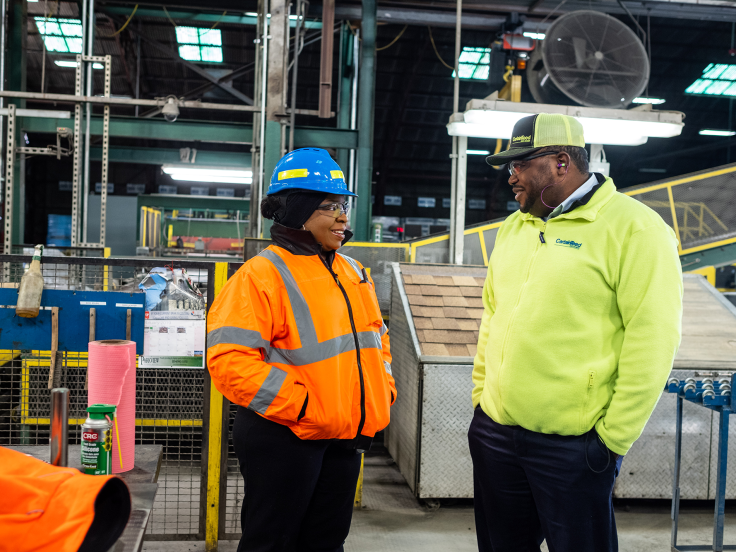A Black man and a Black woman are standing in a factory setting. She wears a blue hard hat and orange safety jacket. He wears a yellow safety jacket. 