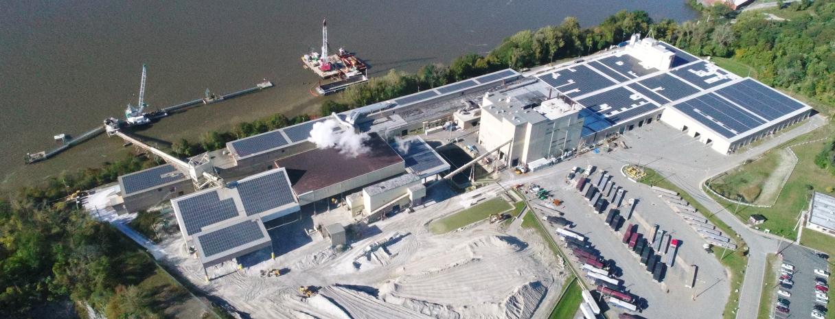 A manufacturing facility sits by a river. Across the roof are solar panels. 