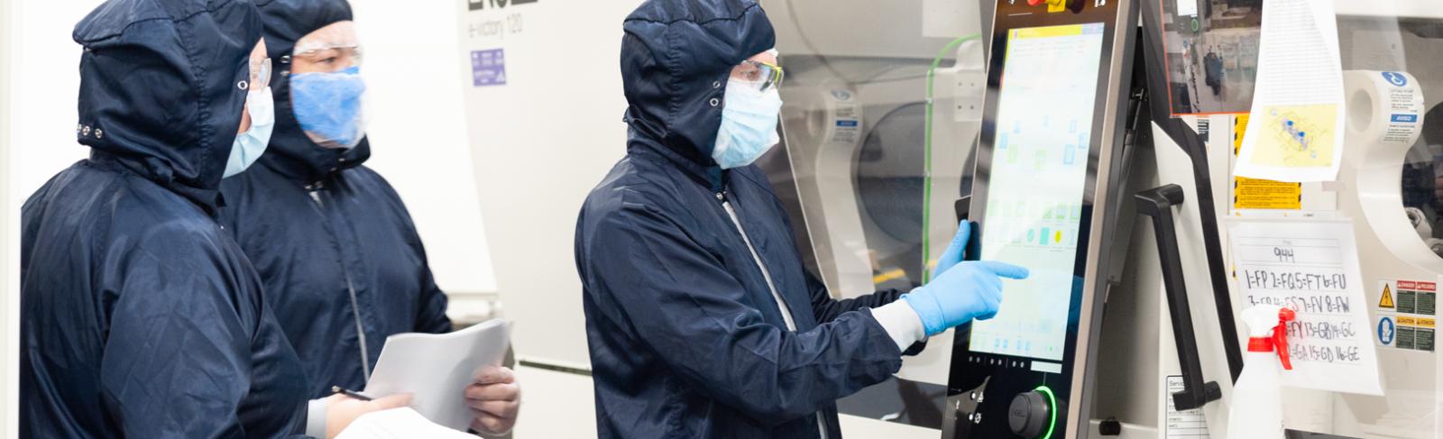 Person in full medical PPE suit using a touch screen attached to a white laboratory machine with two other people in full medical PPE taking notes.