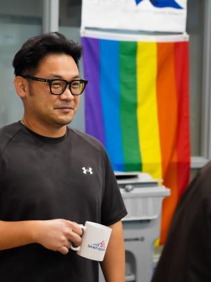 An Asian man stands in an office speaking with a colleague while holding a coffee mug. Behind him hangs a rainbow flag.