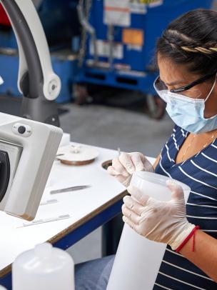woman wearing a surgical mask, gloves and protective eyewear holding a plastic cylinder at a work bench.