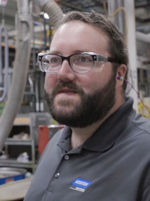 A man wearing safety goggles and a Norton polo shirt stands on the manufacturing floor