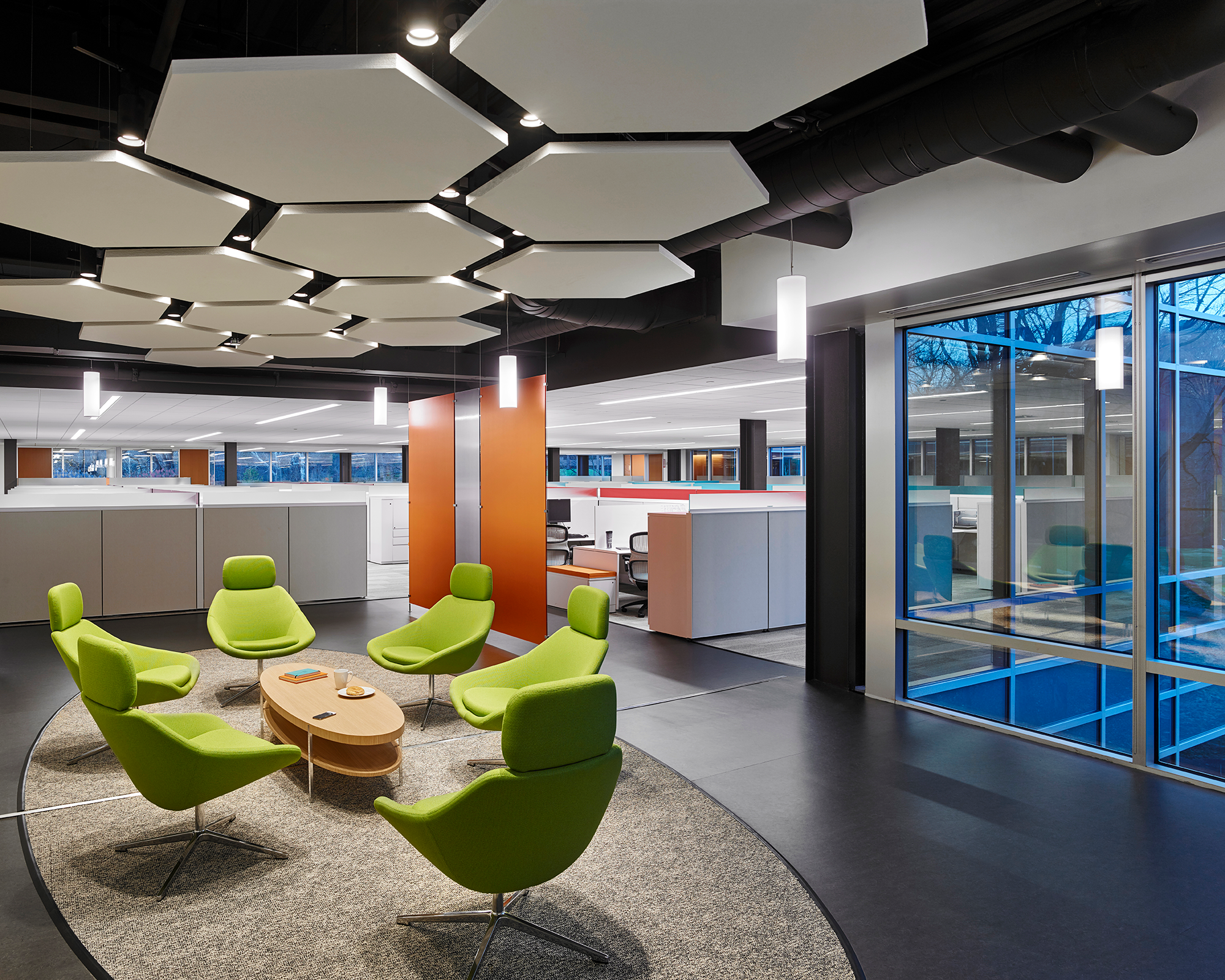 Hexagonal white ceiling panels hang above a seating area with four green chairs around a coffee table. There are cubicles in the background. 