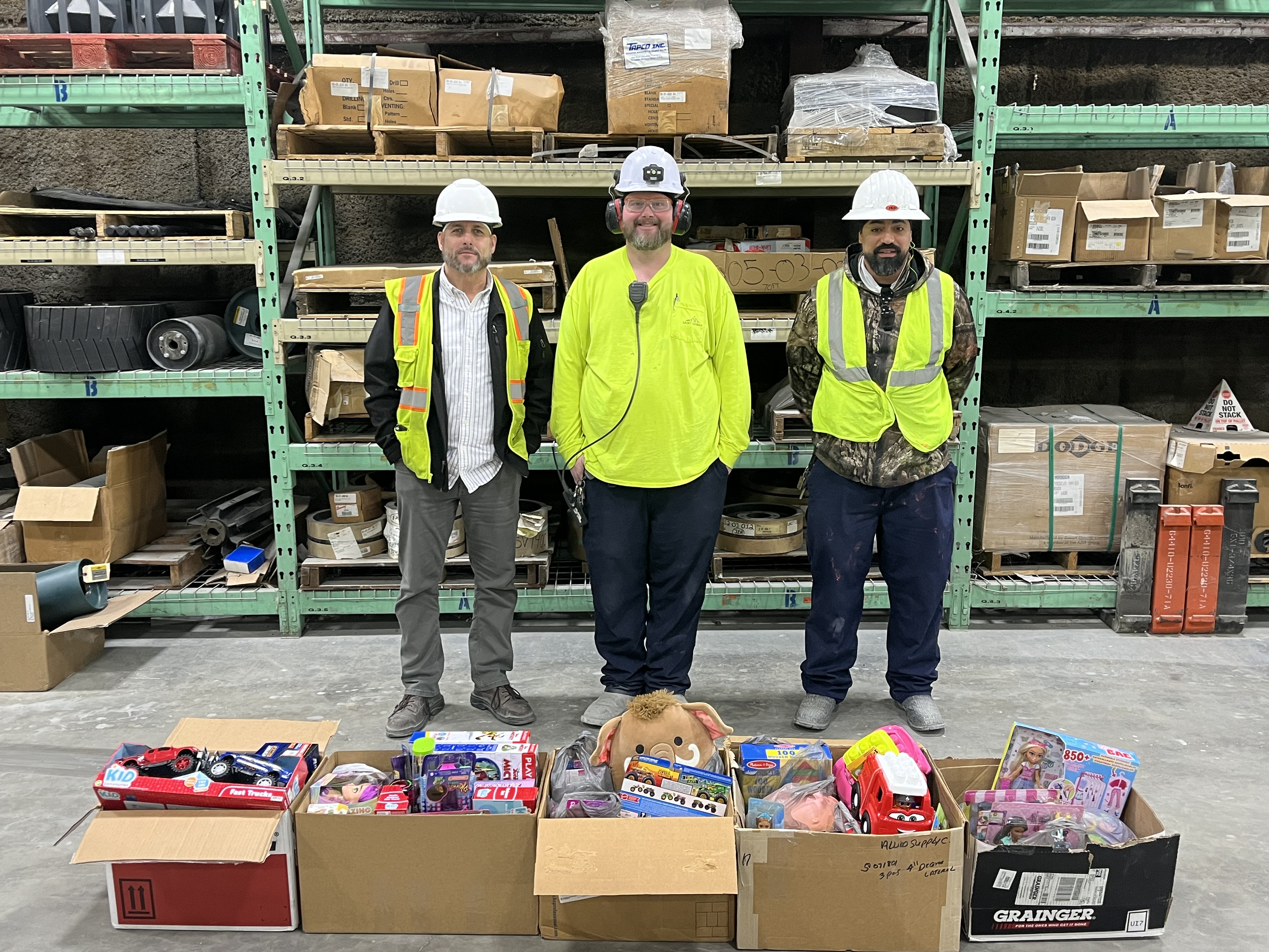 Three men in safety gear stand in a warehouse in front of several cardboard boxes filled with toys