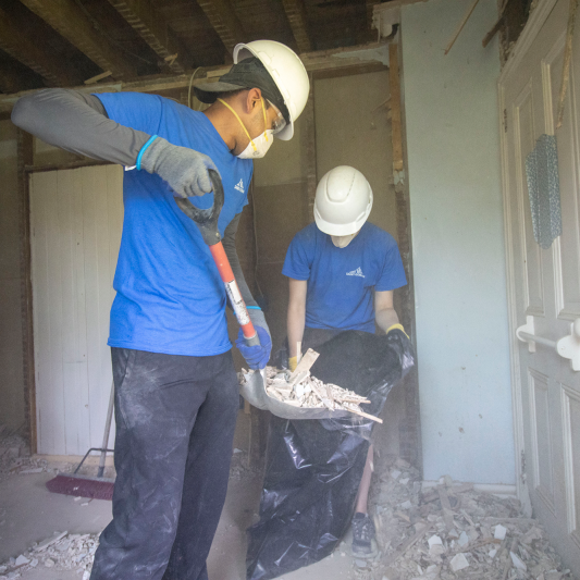 Two people wearing matching shirts are cleaning up after a demolition. One person is holding a trash bag while the other is shoveling debris into it. 