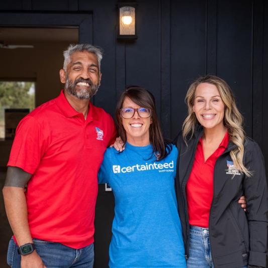 A man wearing a red shirt and his wife, also wearing a white shirt, pose with a CertainTeed employee in front of their new home