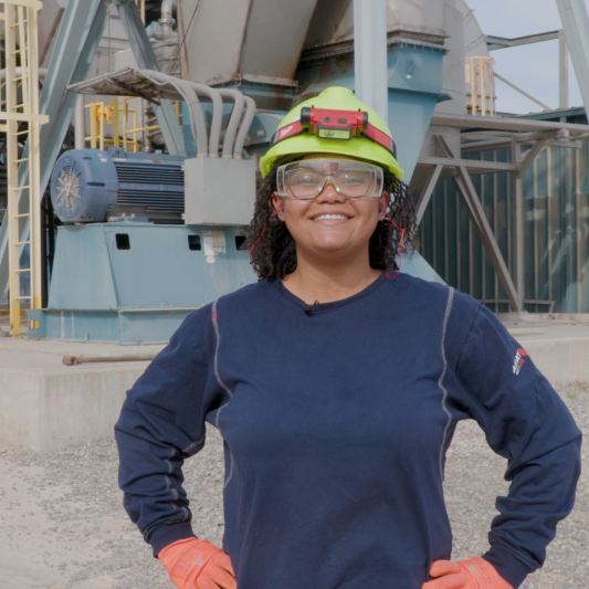 A Black woman wearing a hard hat and safety goggles stands on the plant grounds