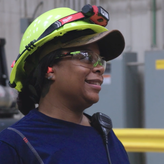 A Black woman with a yellow hard hat is on the manufacturing plant floor