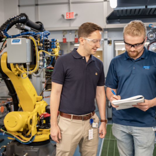 Two engineers at Saint-Gobain North America are discussing notes beside industrial robotic equipment, representing careers in the manufacturing industry.