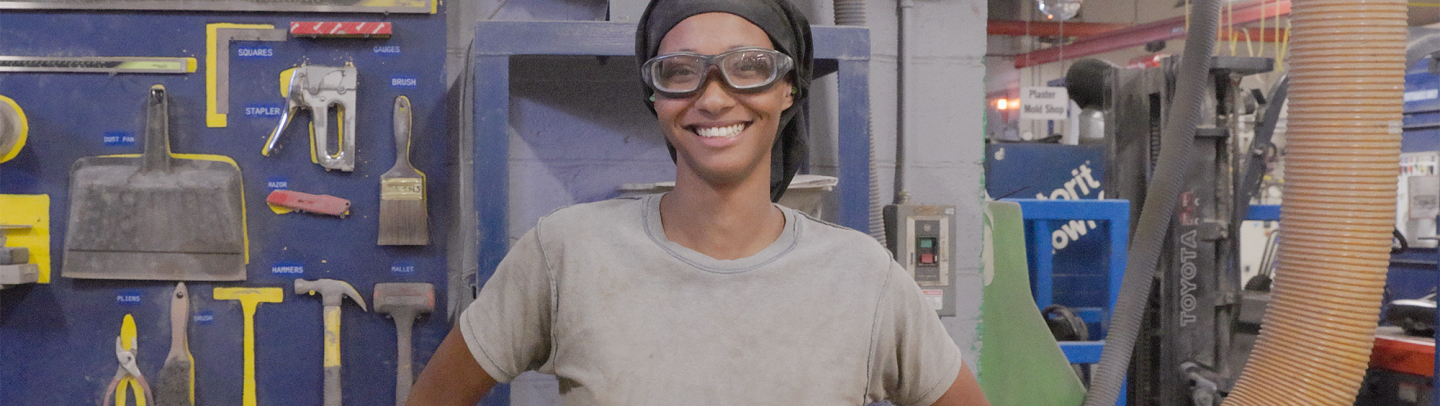 Woman wearing eye protection stands on the ceramics plant floor in front of the tool wall 