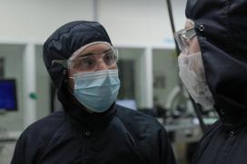 A man in a blue safety suit that covers his entire head and body, safety goggles, and a face mask speaks to a coworker also in safety gear.