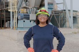 A Black woman wearing a hard hat and safety goggles stands on the plant grounds