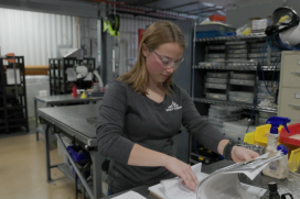 A young blonde women wearing safety gear and a Saint-Gobain shirt works in her plant