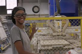 A Black woman wearing safety goggles is standing near a yellow chain link fence inside of a ceramics plant