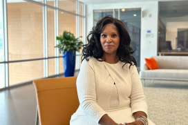 A black woman wearing a white dress sits in an office building lobby area. The walls behind her are glass.