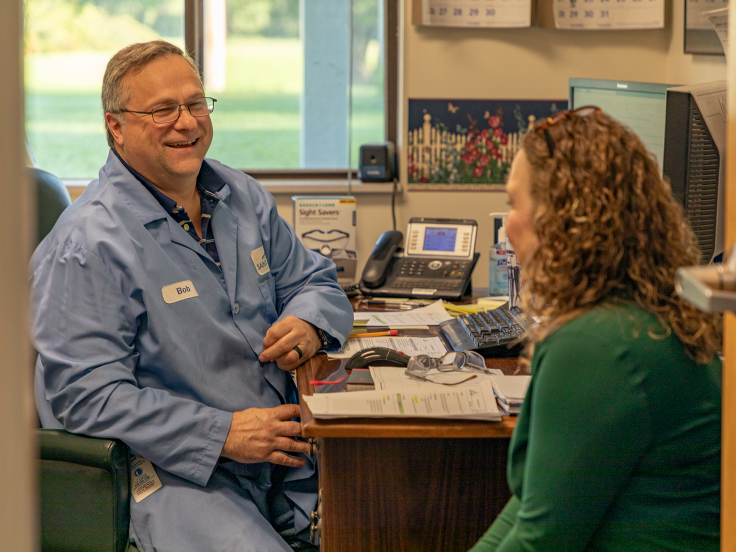 A man sits at his desk while speaking with a female coworker