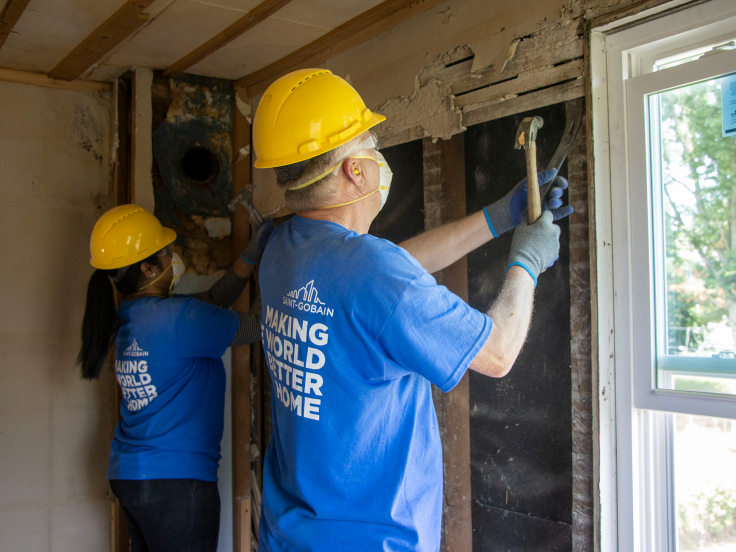 Two people are working on a home building by a window, wearing yellow hard hats and blue tshirts that say "Making The World A Better Home"