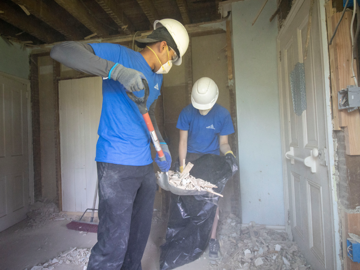 Two people wearing matching shirts are cleaning up after a demolition. One person is holding a trash bag while the other is shoveling debris into it. 