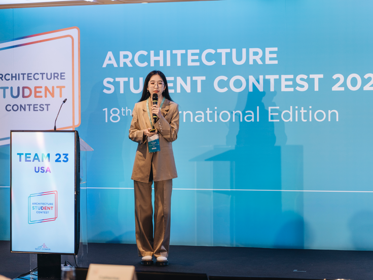 A young woman stands in front of a backdrop that says "Architecture Student Contest 2023" and the podium next to her says USA.