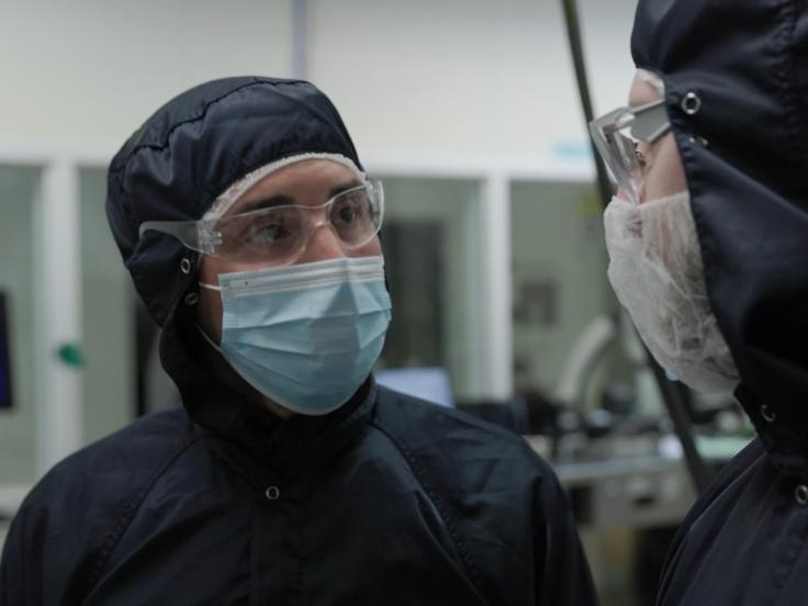 A man in a blue safety suit that covers his entire head and body, safety goggles, and a face mask speaks to a coworker also in safety gear.