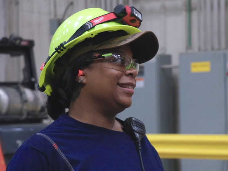 A Black woman with a yellow hard hat is on the manufacturing plant floor