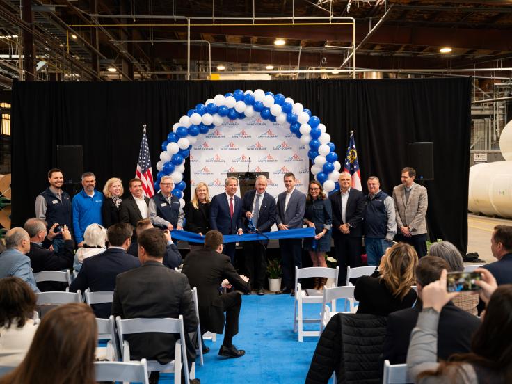 14 people wearing suits stand behind a blue ribbon. The man in the center, Mark Rayfield, holds large scissors on the ribbon. They are standing on the plant floor of CertainTeed's Peachtree City, GA roofing facility in front of a Saint-Gobain banner. 