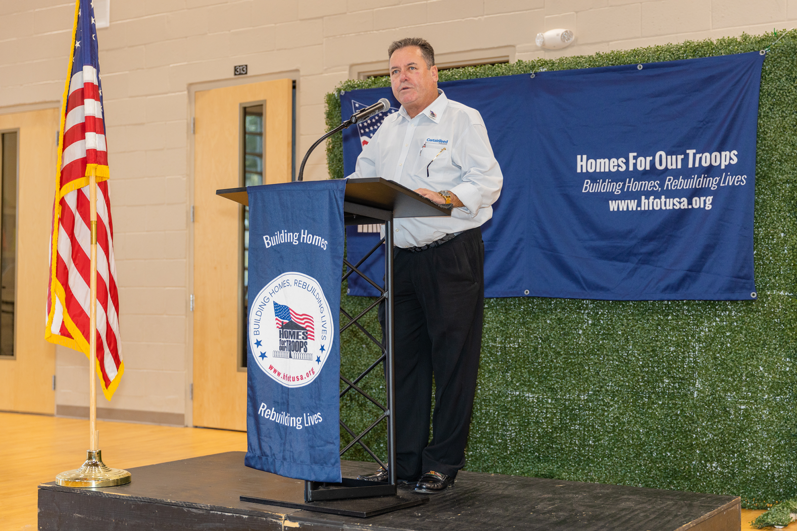 A CertainTeed employee is speaking at a Homes For Our Troops podium at an event.