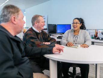 Two men and one woman seated around a round table in an office setting 