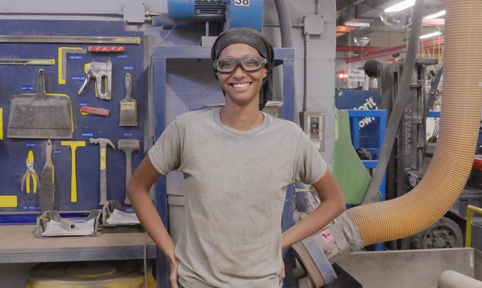 A Black woman wearing a head covering and safety goggles stands on the manufacturing floor