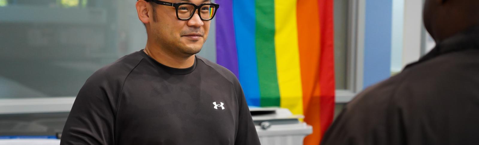 An Asian man stands in an office speaking with a colleague while holding a coffee mug. Behind him hangs a rainbow flag.