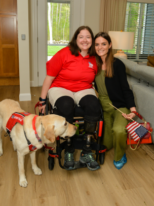 A Golden Retriever service dog sits next to its owner, a woman military veteran in a wheelchair. Another woman stands beside her holding an American flag