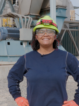 A Black woman wearing a hard hat and safety goggles stands on the plant grounds