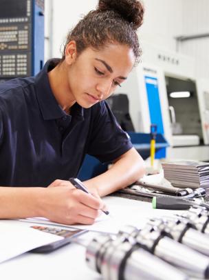 Young female design engineer working with blueprints and tools on her desk, there is manufacturing machinery and a prototyping worker in the background