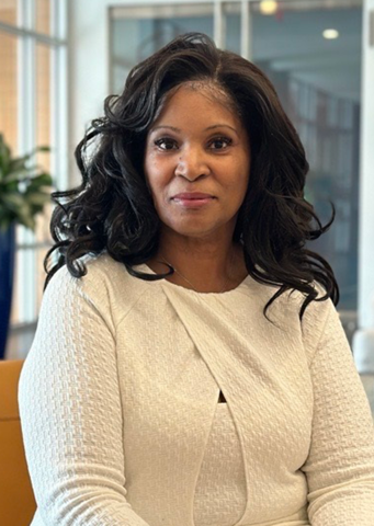 Black woman wearing a white dress sits in an office lobby. The walls behind her are glass. 